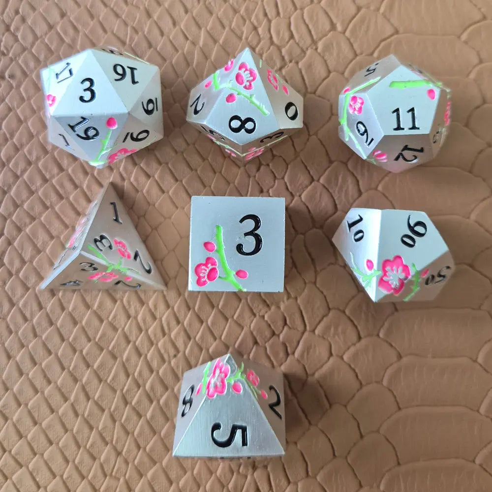 A set of silver metal dice with pink flowers and green stems and black numbers engraved, on a beige textured surface