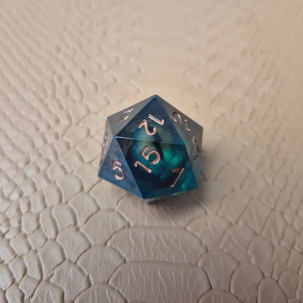 A set of clear green tinted liquid core resin dice with moving dragon eye inside and gold numbers engraved, displayed on a beige scale surface