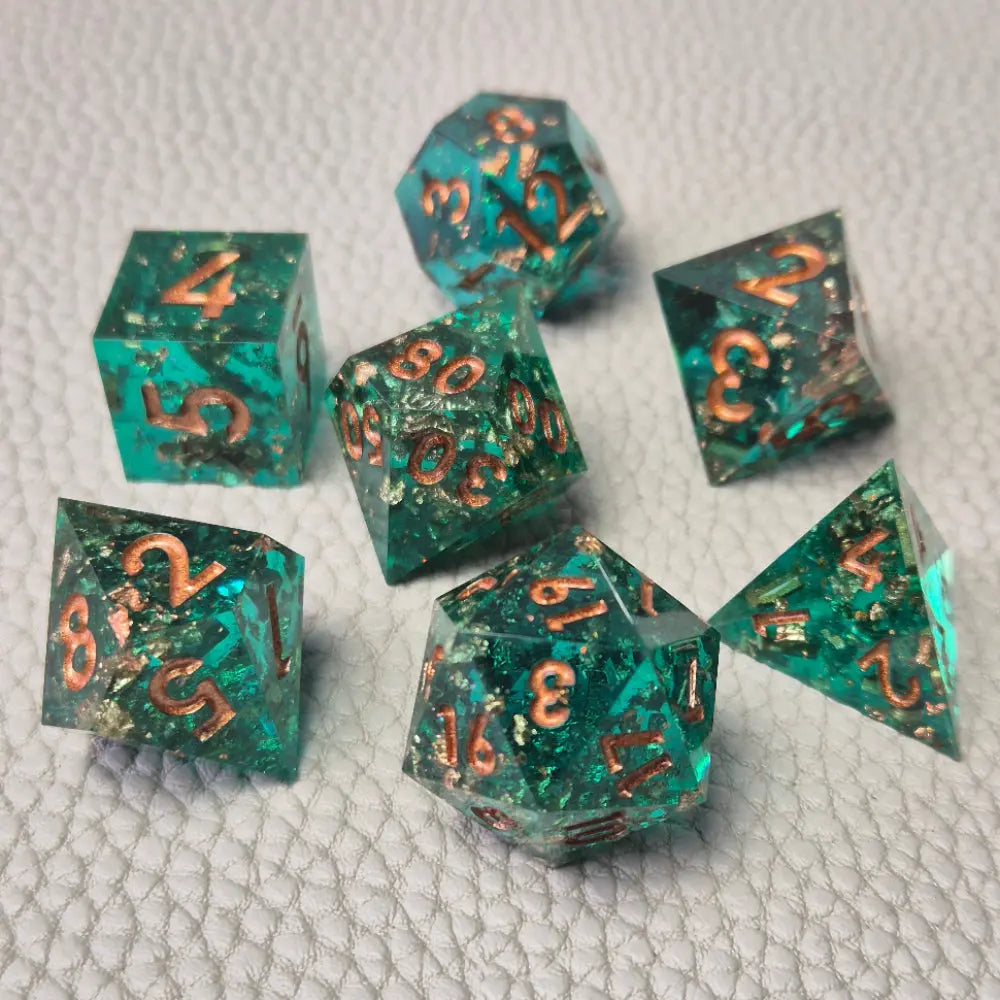 A set of turquoise tinted clear sharp edge resin dice filled with gold flakes. Displayed on a white textured surface