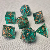 A set of turquoise tinted clear sharp edge resin dice filled with gold flakes. Displayed on a white textured surface