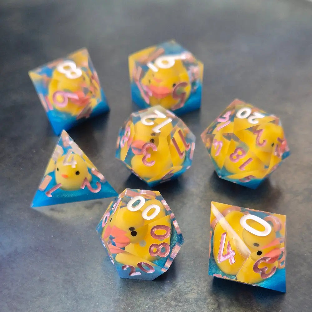 Whimsical sharp-edge dice set with floating yellow ducks and pink numbering, set in clear resin over a blue water-like base.