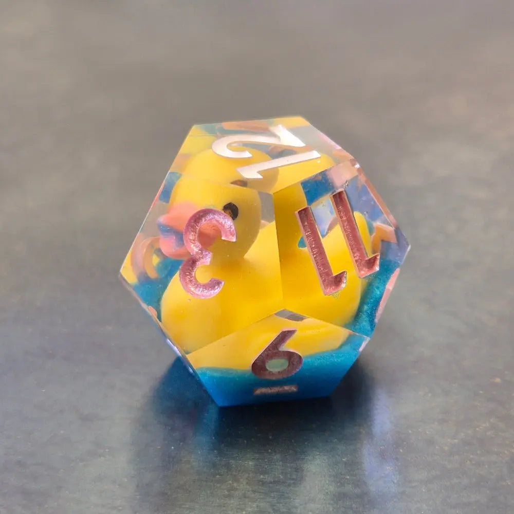 Whimsical sharp-edge D12 with floating yellow ducks and pink numbering, set in clear resin over a blue water-like base.