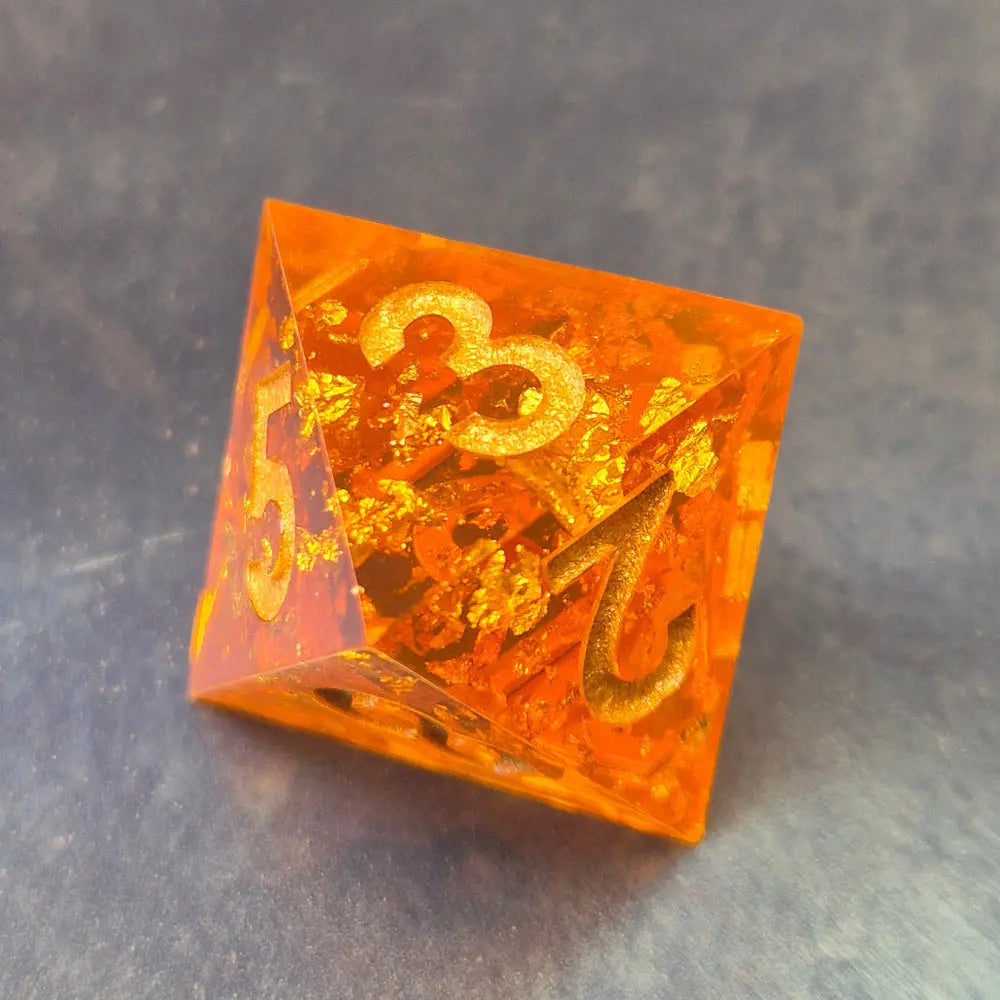 A D8 sharp-edge orange resin dice with sparkling gold foil and gold numbers, displayed on a metallic surface.