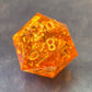 A D20 sharp-edge orange resin dice with sparkling gold foil and gold numbers, displayed on a metallic surface.