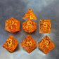 A set of sharp-edge orange resin dice with sparkling gold foil and gold numbers, displayed on a metallic surface.