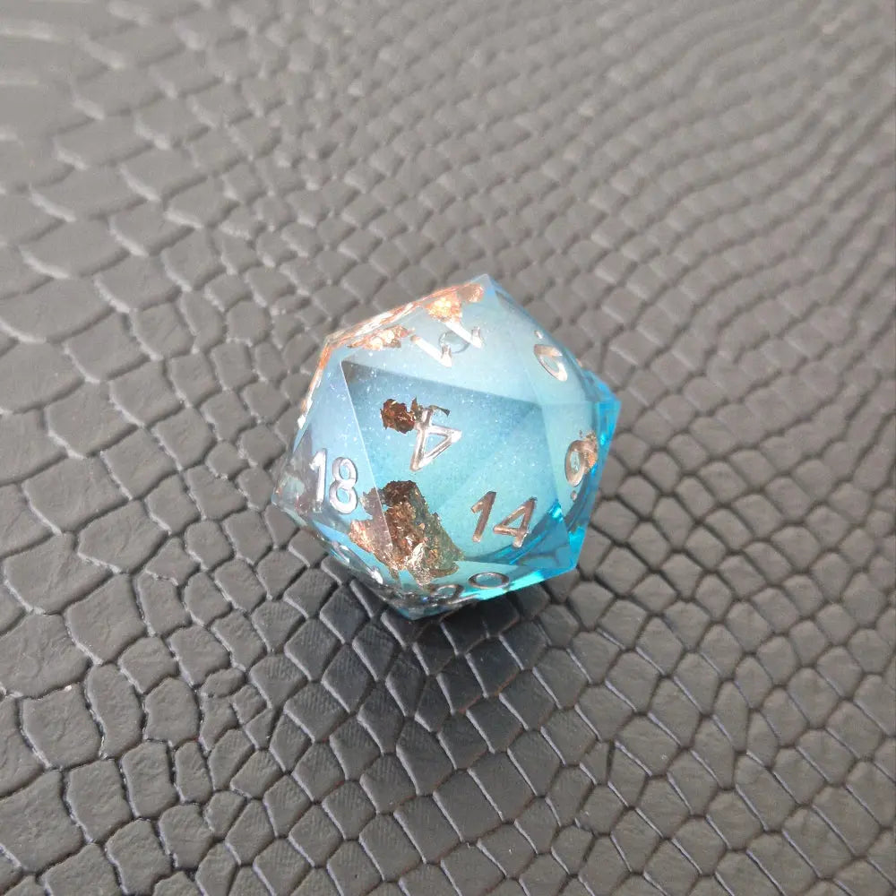A set of clear light blue tinted liquid core dice with floating bronze flakes and white numbers, displayed on a black textured surface