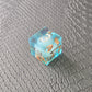 A set of clear light blue tinted liquid core dice with floating bronze flakes and white numbers, displayed on a black textured surface