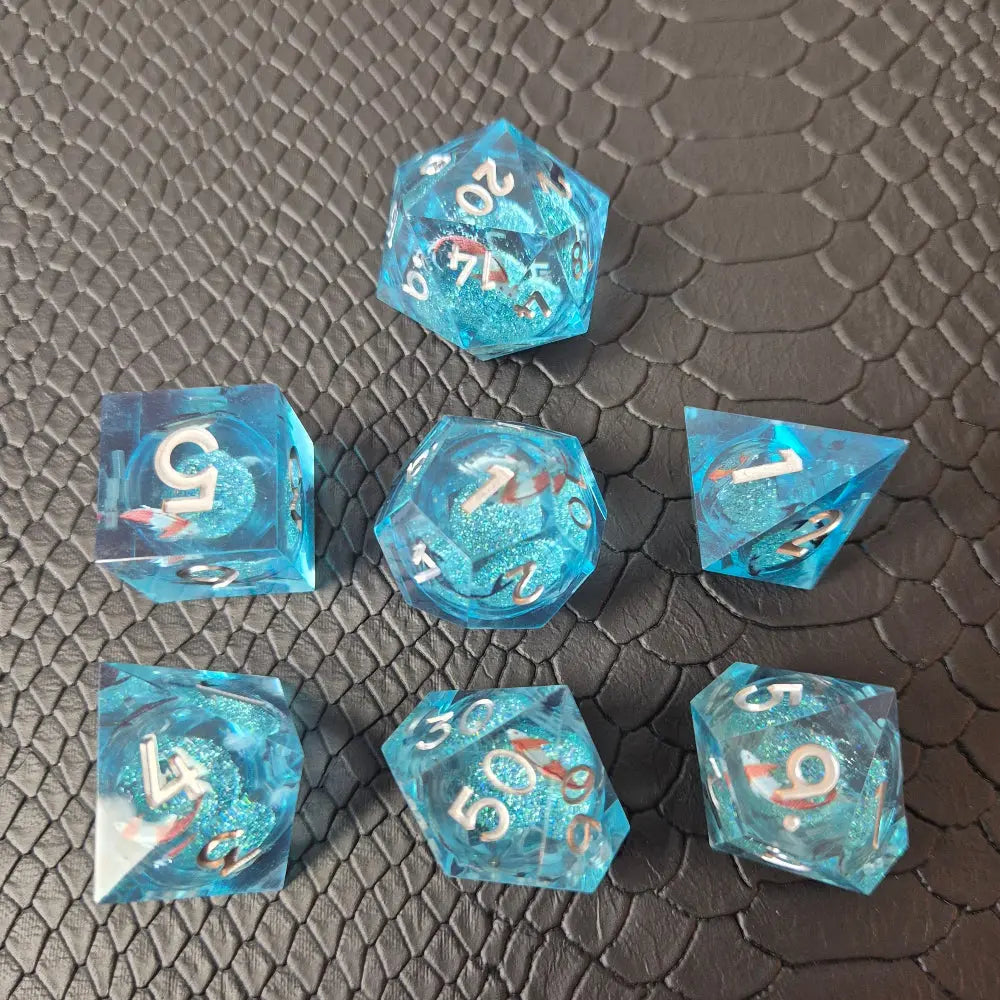 A set of clear light blue tinted liquid core dice with koi fish and glitters inside, displayed on a black textured surface