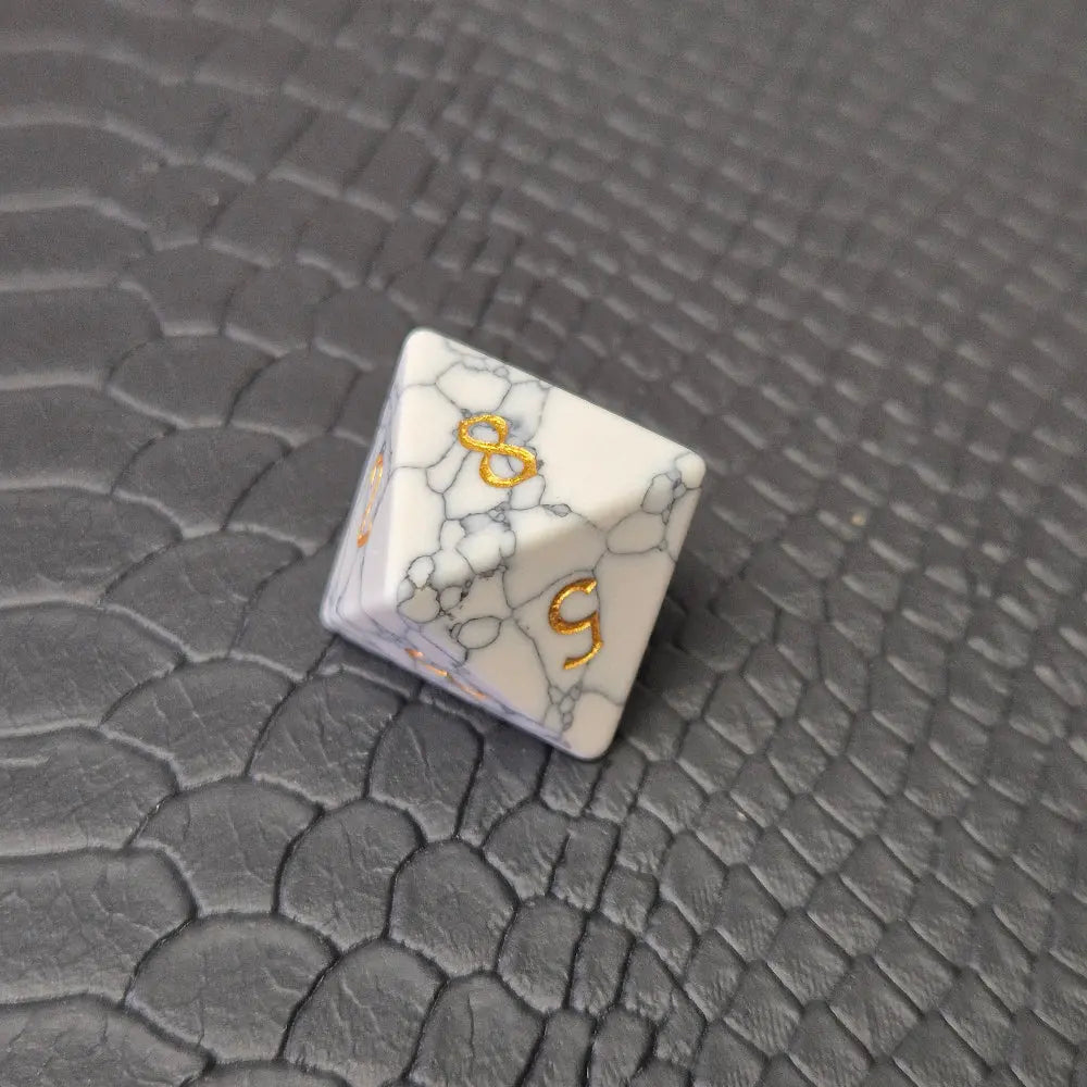 D8 white marble-textured gemstone die with gold numbering, displayed on a black leather surface.