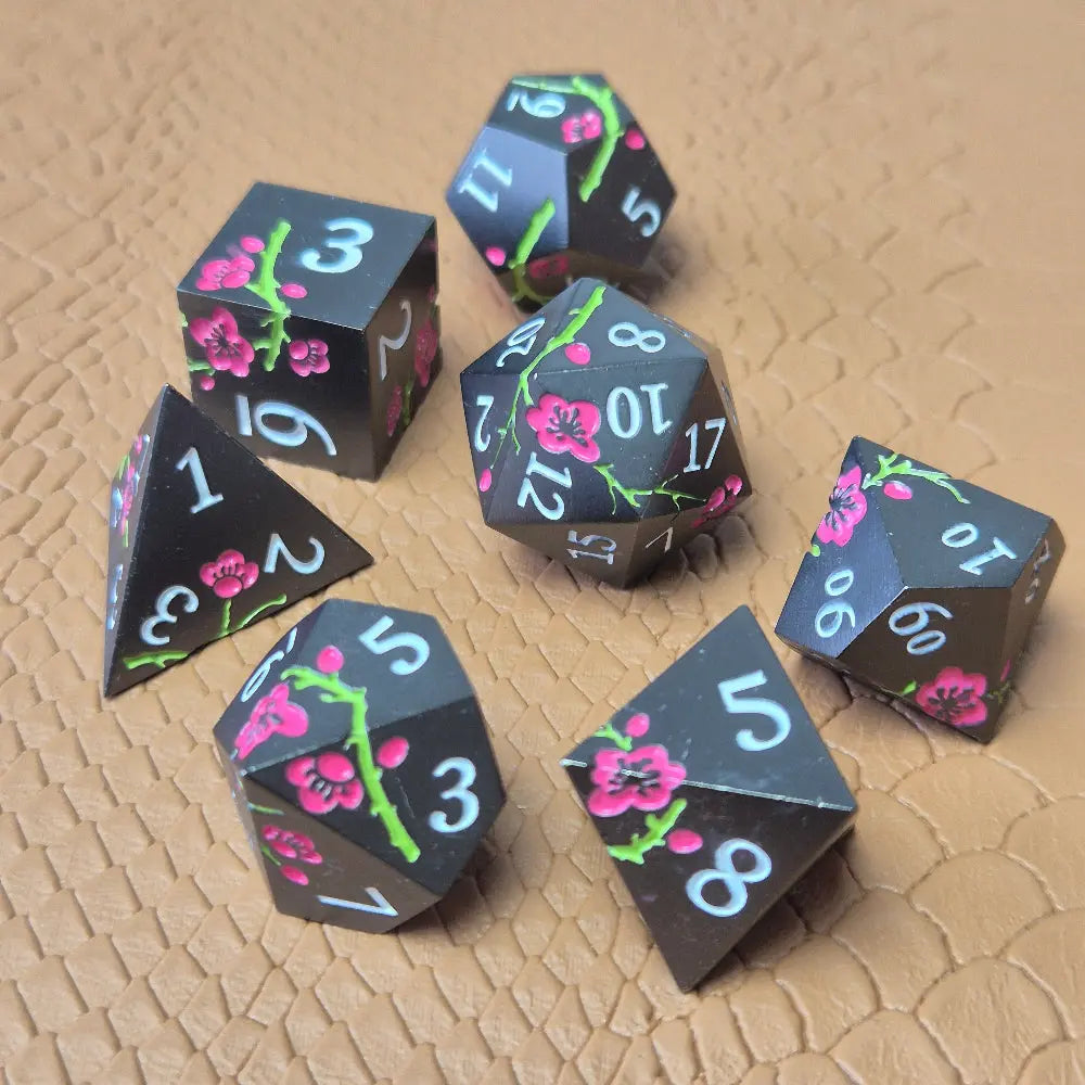 A set of black metal dice with white numbers and pink flowers with green stems, displayed on a brown textured surface.