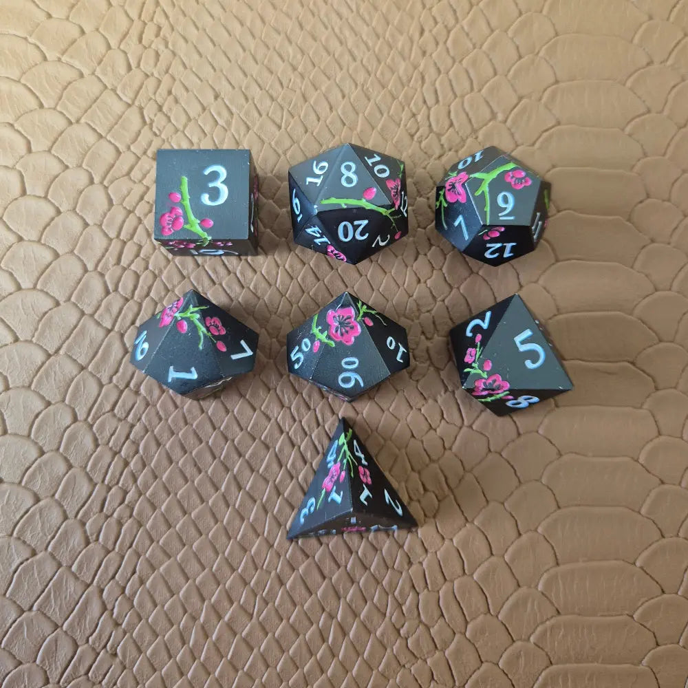 A set of black metal dice with white numbers and pink flowers with green stems, displayed on a brown textured surface.