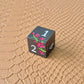 A set of black metal dice with white numbers and pink flowers with green stems, displayed on a brown textured surface.