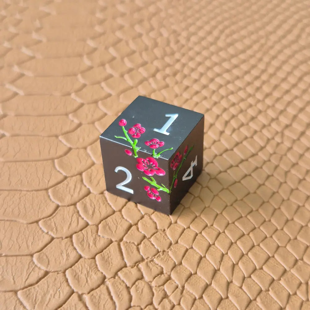 A set of black metal dice with white numbers and pink flowers with green stems, displayed on a brown textured surface.