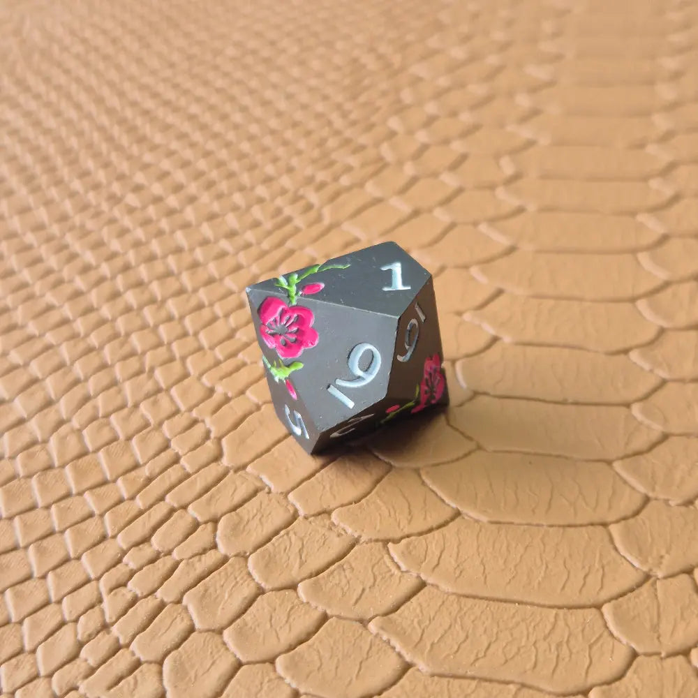 A set of black metal dice with white numbers and pink flowers with green stems, displayed on a brown textured surface.
