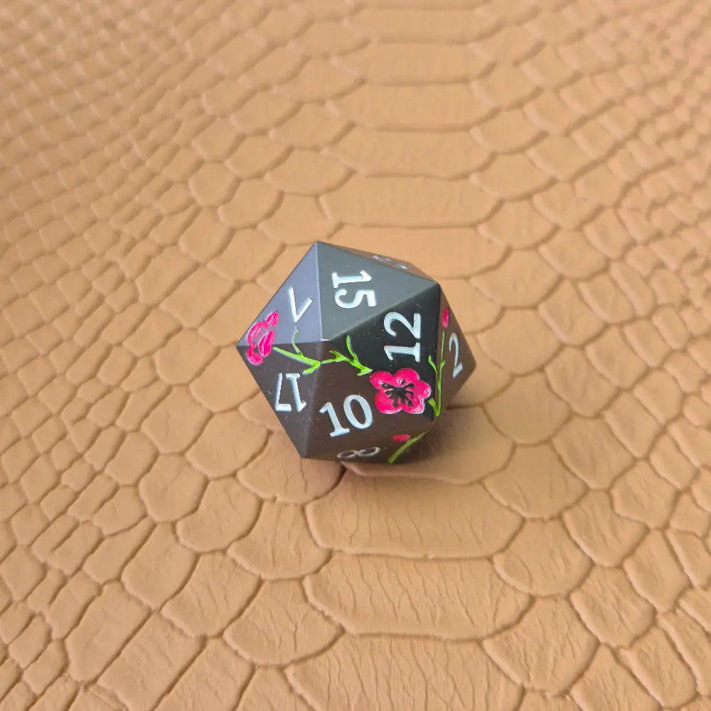 A set of black metal dice with white numbers and pink flowers with green stems, displayed on a brown textured surface.