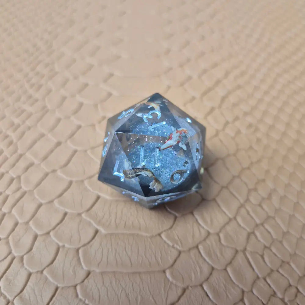 A set of clear grey tinted liquid core resin dice with koi fish inside filled with glitters on a beige textured surface