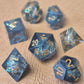 A set of sharp-edge resin dice with ocean-blue transparency, shimmering foil inclusions, and gold numbering, displayed on a textured beige surface.