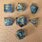 A set of sharp-edge resin dice with ocean-blue transparency, shimmering foil inclusions, and gold numbering, displayed on a textured beige surface.