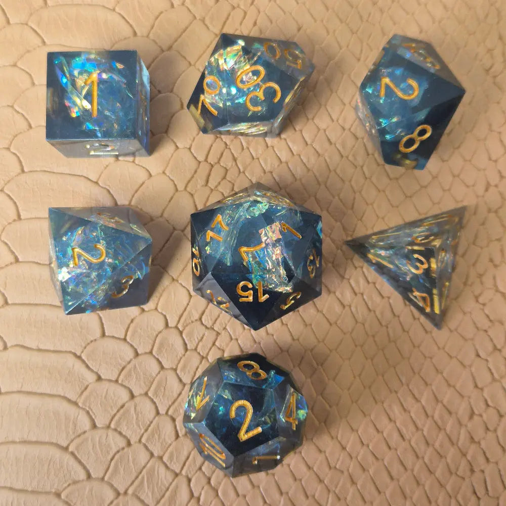 A set of sharp-edge resin dice with ocean-blue transparency, shimmering foil inclusions, and gold numbering, displayed on a textured beige surface.