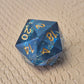 A D20 sharp-edge resin dice with ocean-blue transparency, shimmering foil inclusions, and gold numbering, displayed on a textured beige surface.