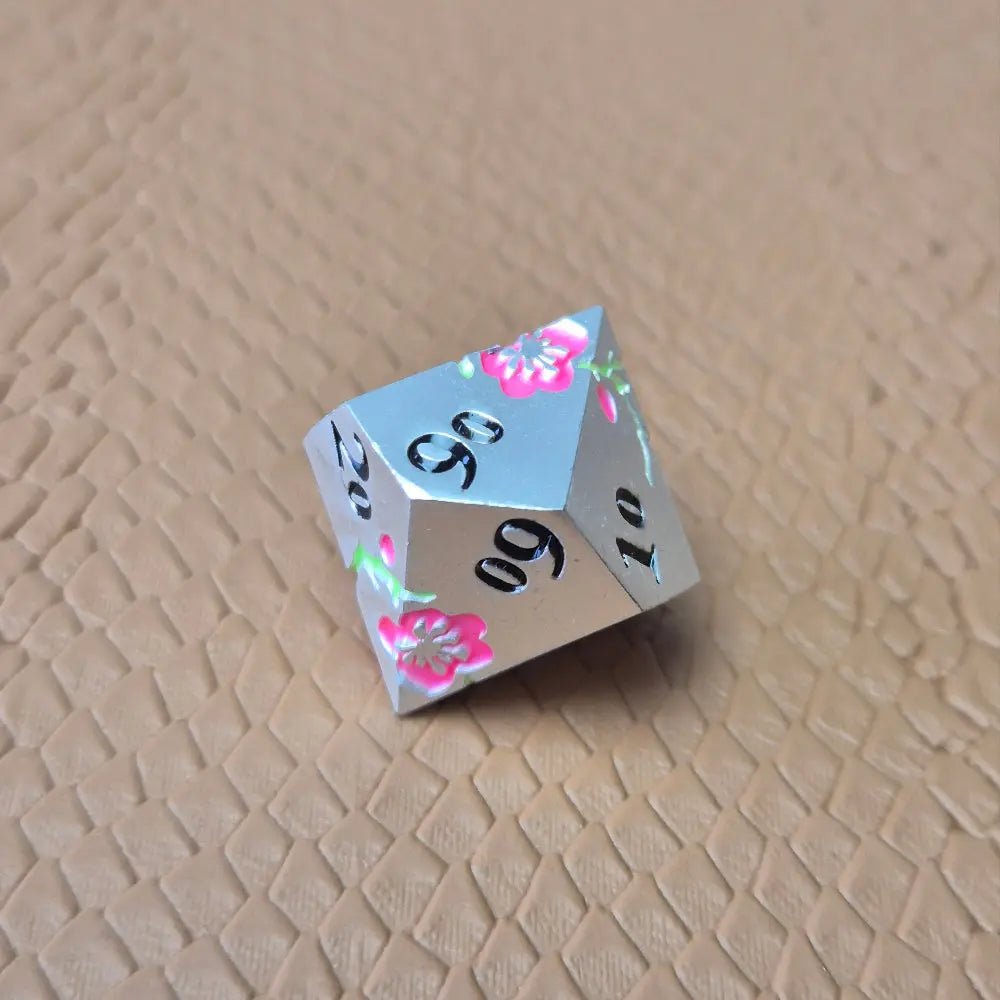 A set of silver metal dice with pink flowers and green stems and black numbers engraved, on a beige textured surface
