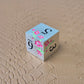 A set of silver metal dice with pink flowers and green stems and black numbers engraved, on a beige textured surface