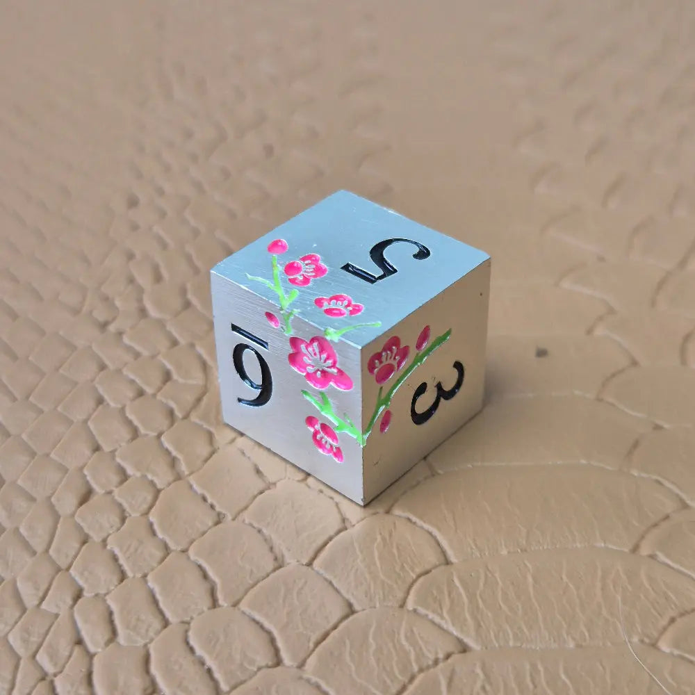 A set of silver metal dice with pink flowers and green stems and black numbers engraved, on a beige textured surface