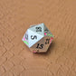 A set of silver metal dice with pink flowers and green stems and black numbers engraved, on a beige textured surface