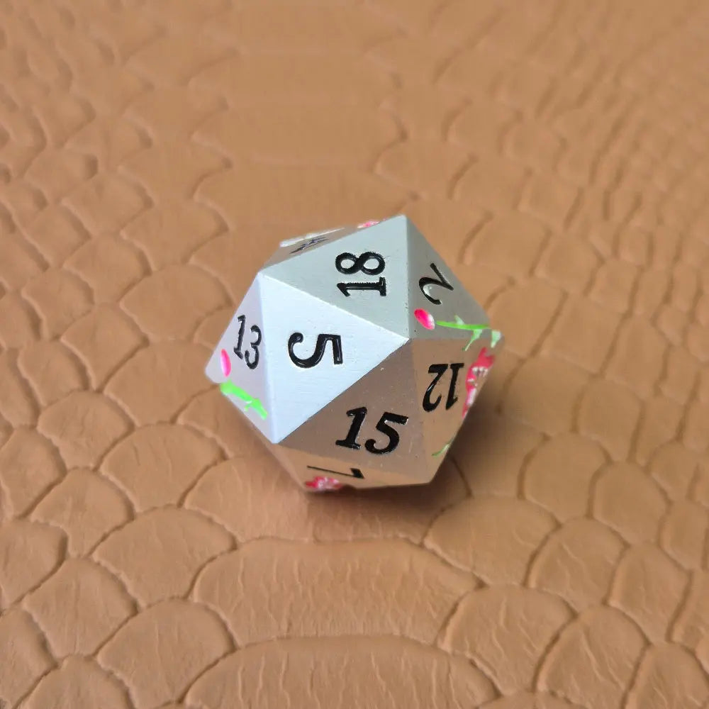 A set of silver metal dice with pink flowers and green stems and black numbers engraved, on a beige textured surface