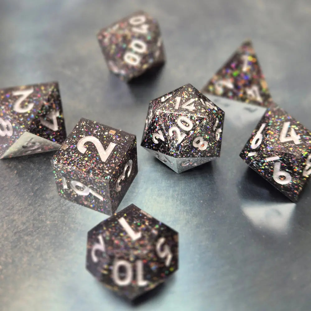 A set of black sharp edge resin dice with white numbers and colourfull sparkles on a blue metal surface