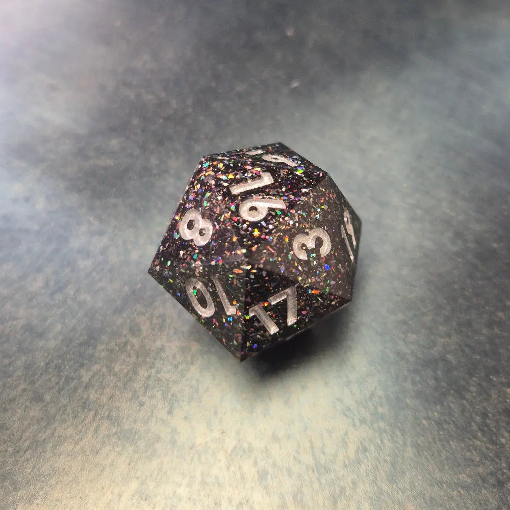 A set of black sharp edge resin dice with white numbers and colourfull sparkles on a blue metal surface