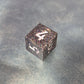 A set of black sharp edge resin dice with white numbers and colourfull sparkles on a blue metal surface