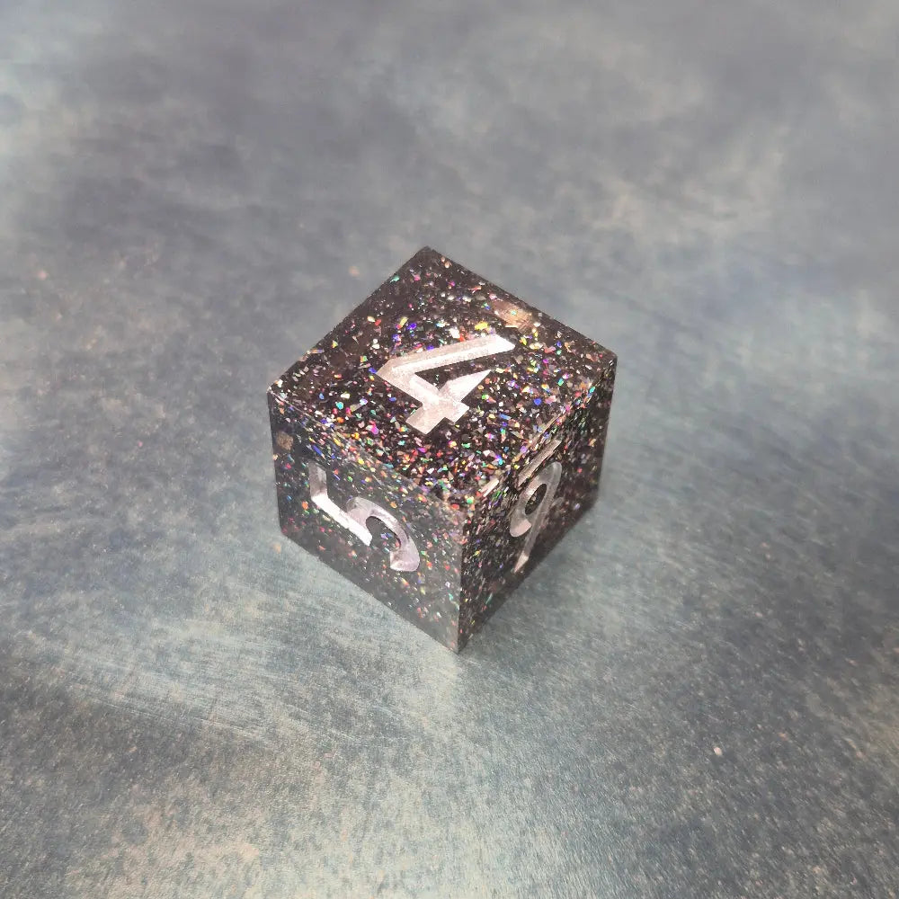 A set of black sharp edge resin dice with white numbers and colourfull sparkles on a blue metal surface