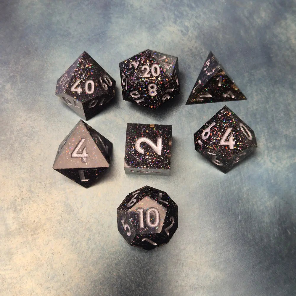 A set of black sharp edge resin dice with white numbers and colourfull sparkles on a blue metal surface