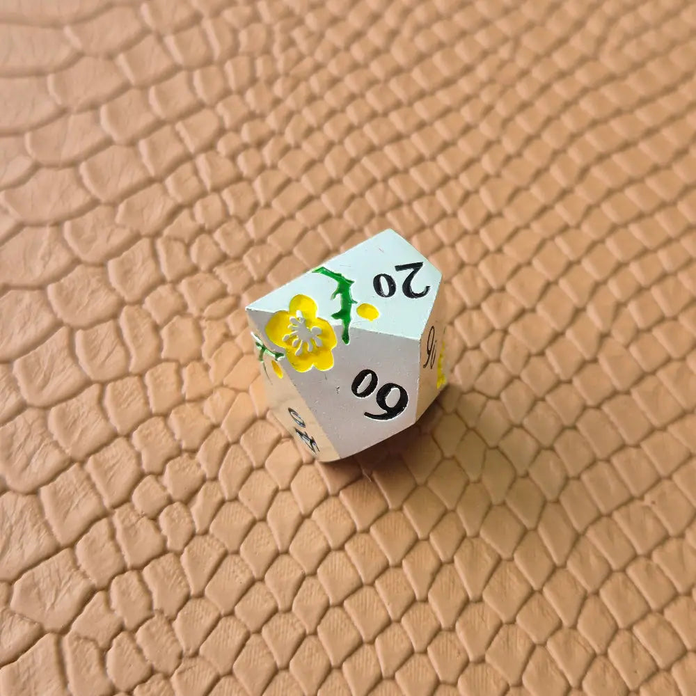 Metal D&D dice set with matte silver finish and painted yellow flowers, displayed on textured leather background.