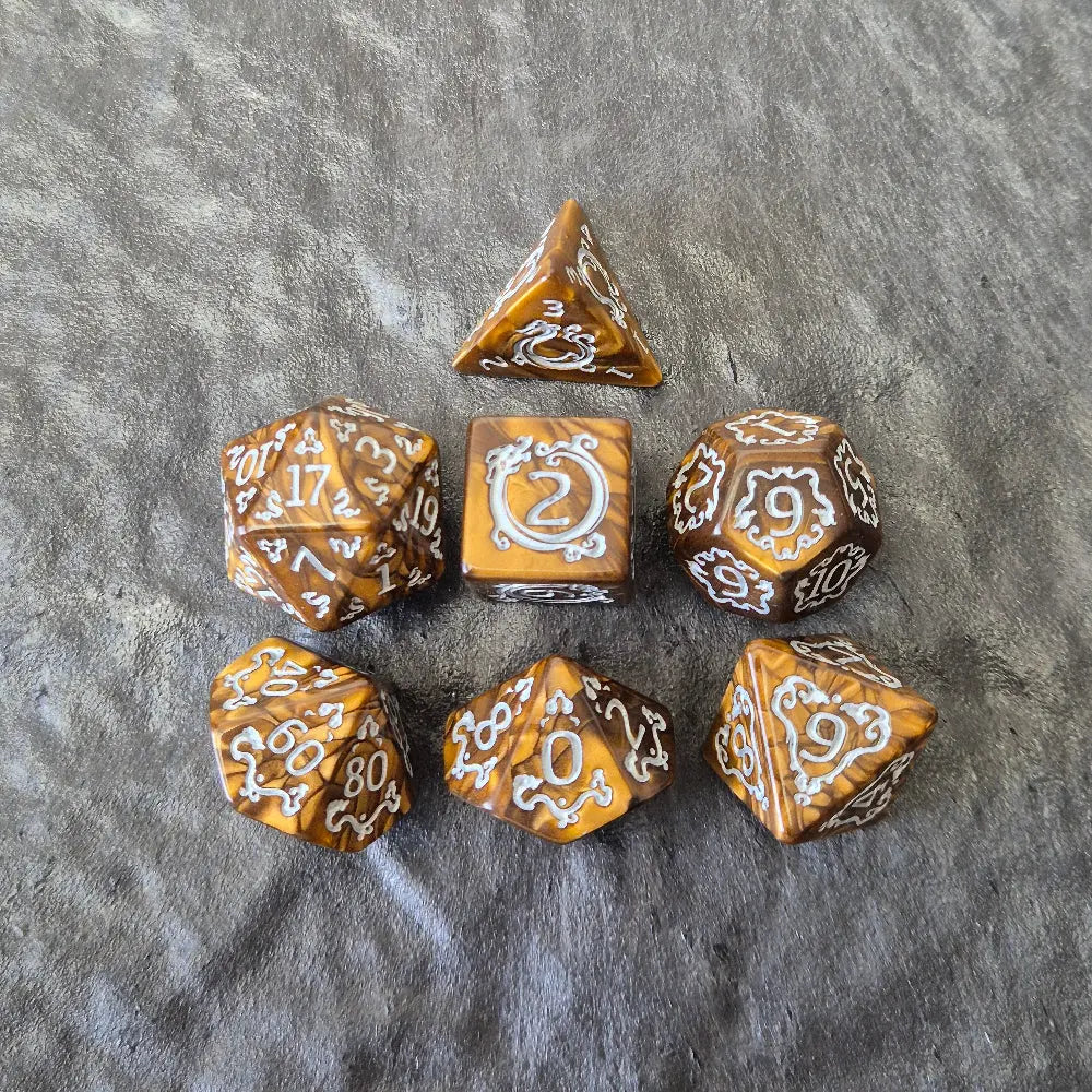 Gold acrylic polyhedral dice set with ornate white fantasy-style numbers displayed on a stone surface