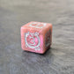Pink acrylic D6 with white arcane-style numbers and elegant swirls, displayed on a textured grey surface.