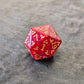 Red acrylic D20 with golden arcane-style numbers and swirling patterns, displayed on a textured grey background.
