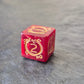 Red acrylic D6 with golden arcane-style numbers and swirling patterns, displayed on a textured grey background.