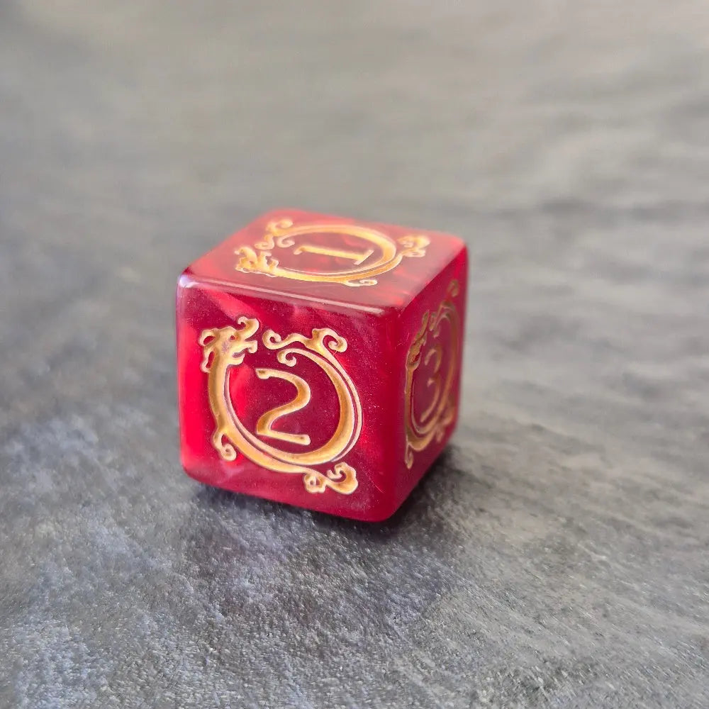 Red acrylic D6 with golden arcane-style numbers and swirling patterns, displayed on a textured grey background.
