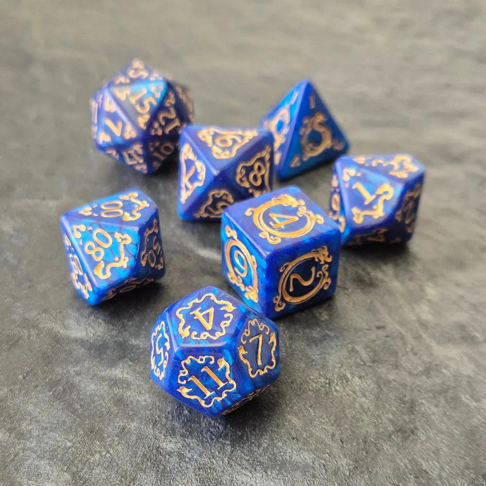 7-piece blue acrylic dice set with ornate gold numbering and arcane motifs, displayed on a dark stone surface.