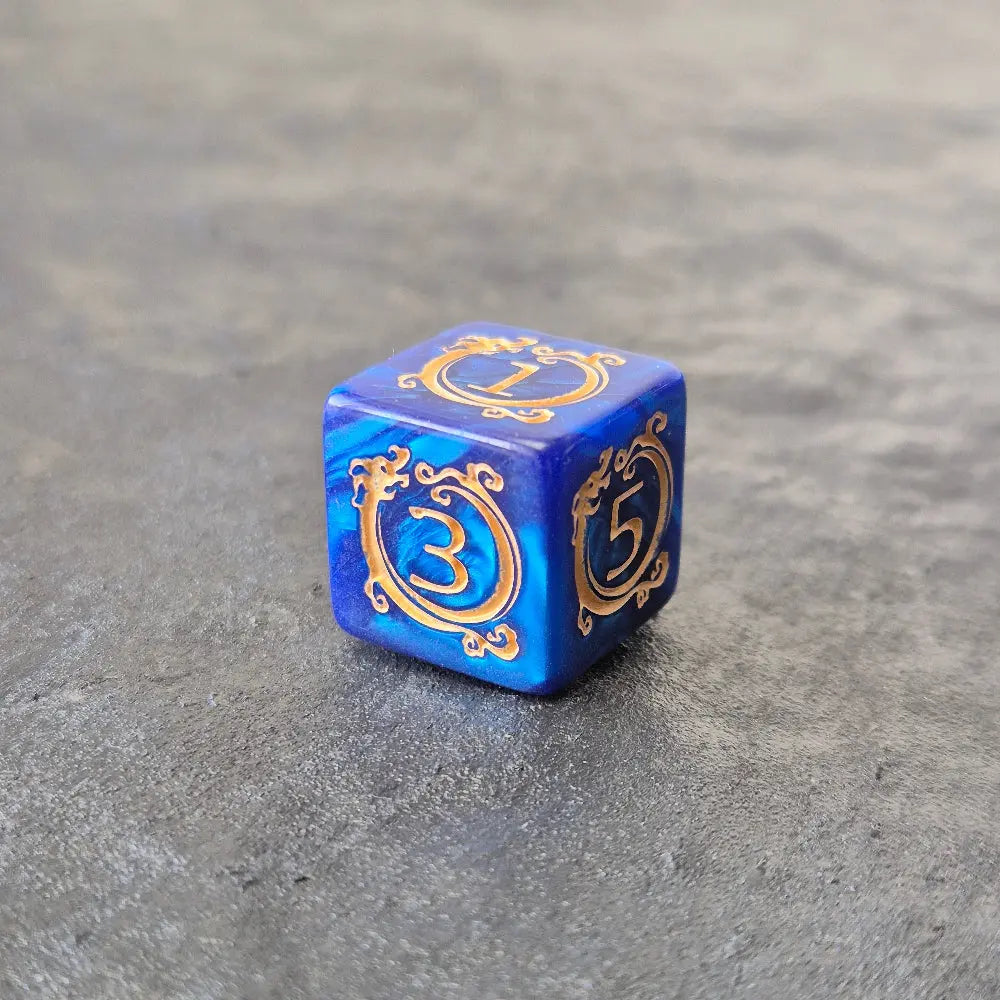D6 blue acrylic dice with ornate gold numbering and arcane motifs, displayed on a dark stone surface.