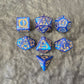 7-piece blue acrylic dice set with ornate gold numbering and arcane motifs, displayed on a dark stone surface.