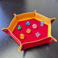 Foldable hexagon-shaped dice tray in yellow & red shown open and filled with colorful polyhedral dice.