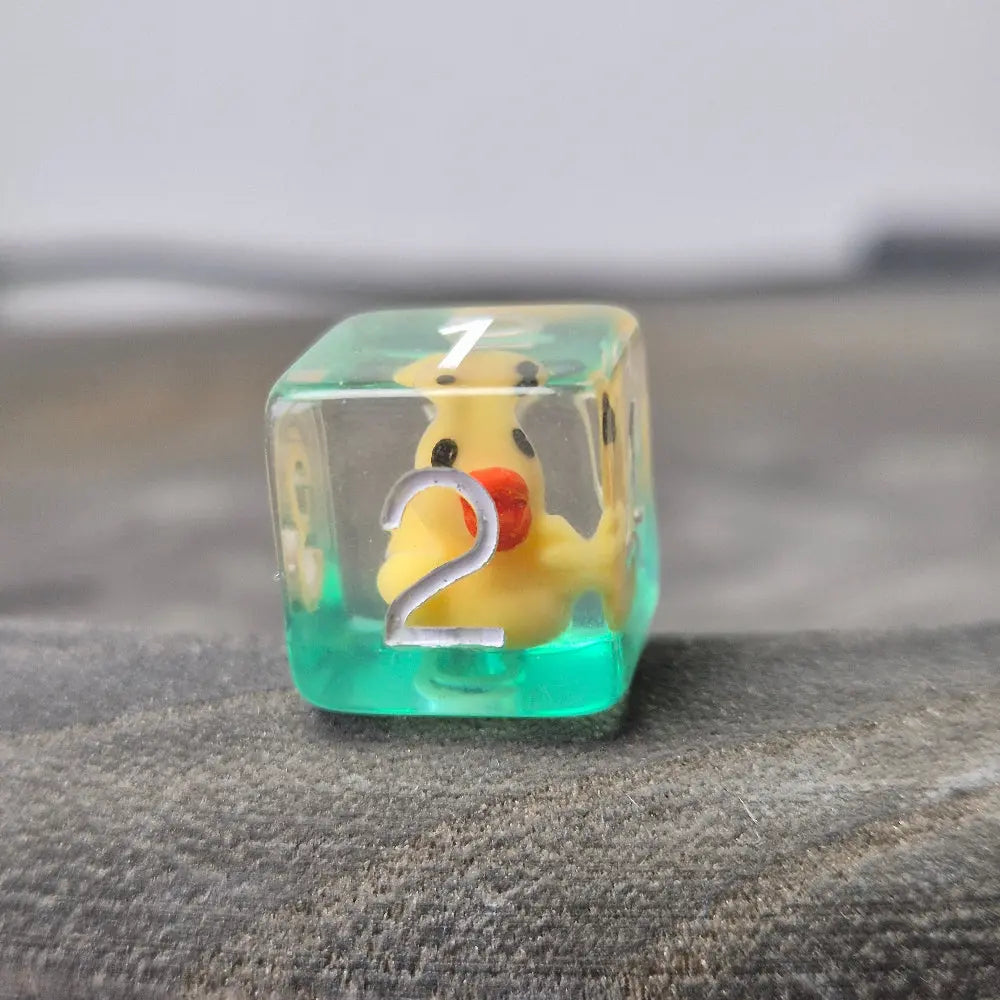 Close-up of transparent D6 resin dice with tiny yellow ducks inside, placed on a textured stone surface.