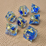 Sharp-edge polyhedral dice set with golden ring embedded in clear and blue resin, gold inked numbers, displayed on a fantasy-style textured surface.