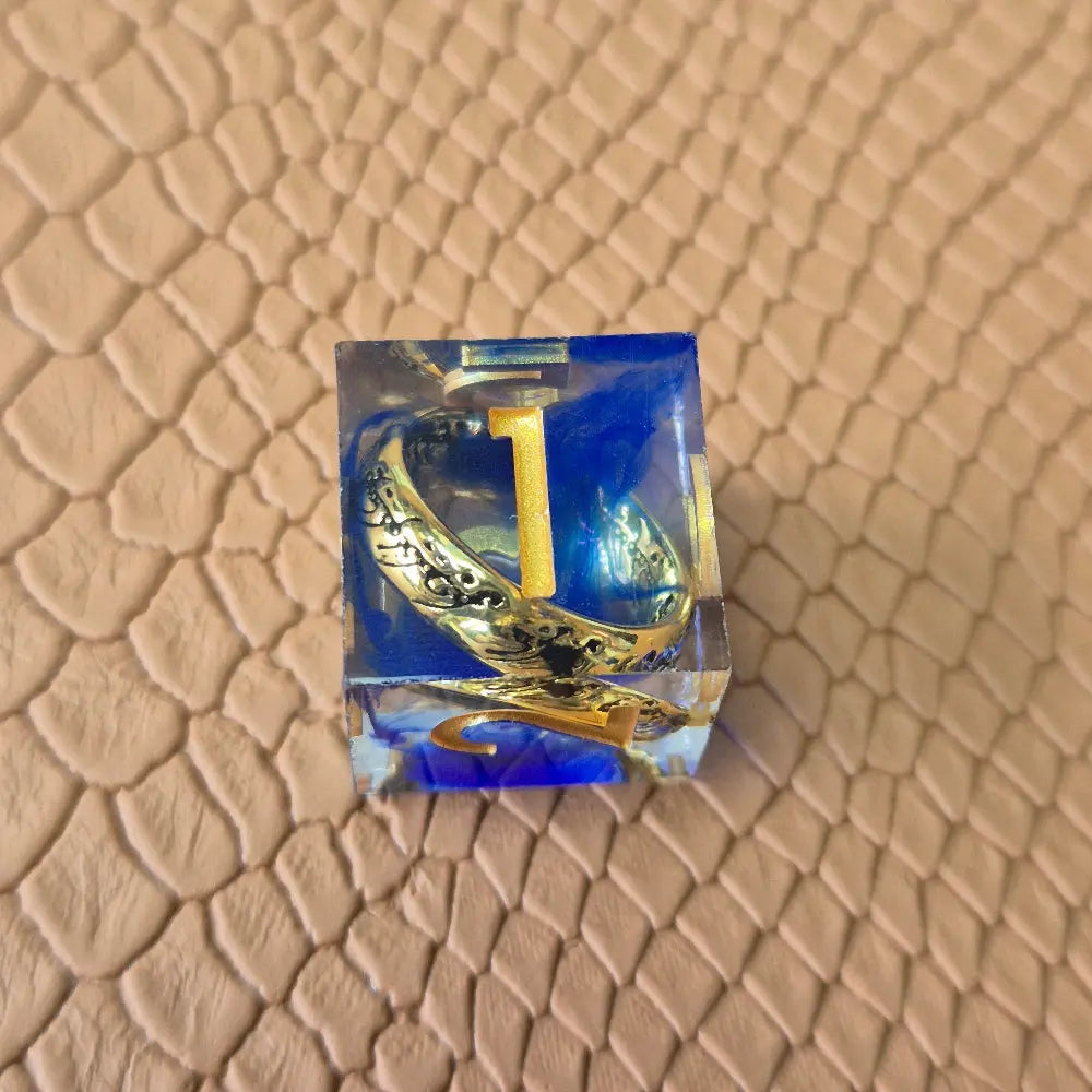 Sharp-edge polyhedral D6 with golden ring embedded in clear and blue resin, gold inked numbers, displayed on a fantasy-style textured surface.