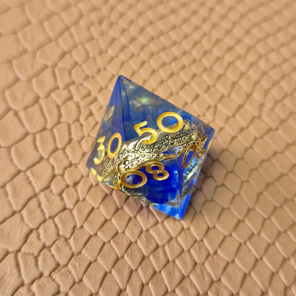 Sharp-edge polyhedral D100 with golden ring embedded in clear and blue resin, gold inked numbers, displayed on a fantasy-style textured surface.