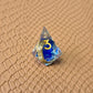 Sharp-edge polyhedral dice set with golden ring embedded in clear and blue resin, gold inked numbers, displayed on a fantasy-style textured surface.