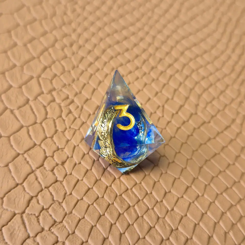 Sharp-edge polyhedral dice set with golden ring embedded in clear and blue resin, gold inked numbers, displayed on a fantasy-style textured surface.